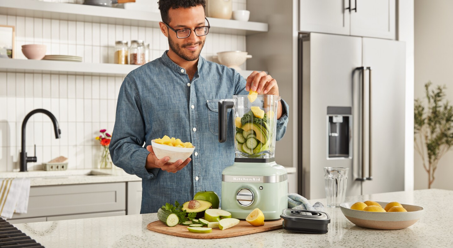 A man adding vegetables to a KitchenAid® blender in a modern kitchen. A man adding vegetables to a KitchenAid® blender in a modern kitchen.