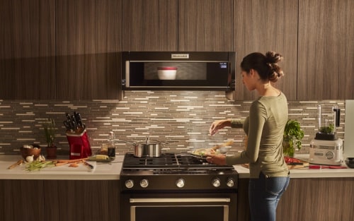 Person preparing a recipe on a gas stovetop below a KitchenAid® low profile microwave